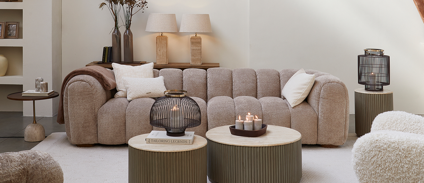 Eine Frau liest auf einem cremefarbenen Bett vor einem großen Rundbogenfenster, umgeben von Regalen, Pflanzen und einem Holz­spiegel.