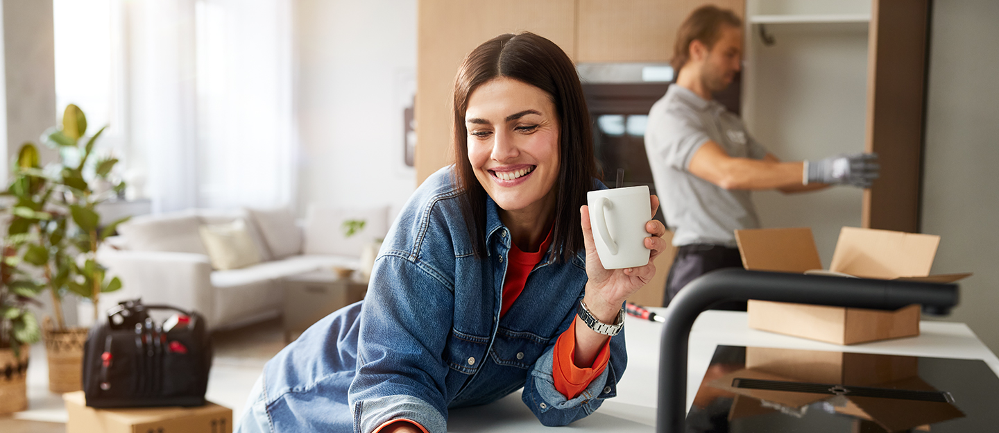 Frau hält eine Tasse, während im Hintergrund ein Monteur Möbel aufbaut.