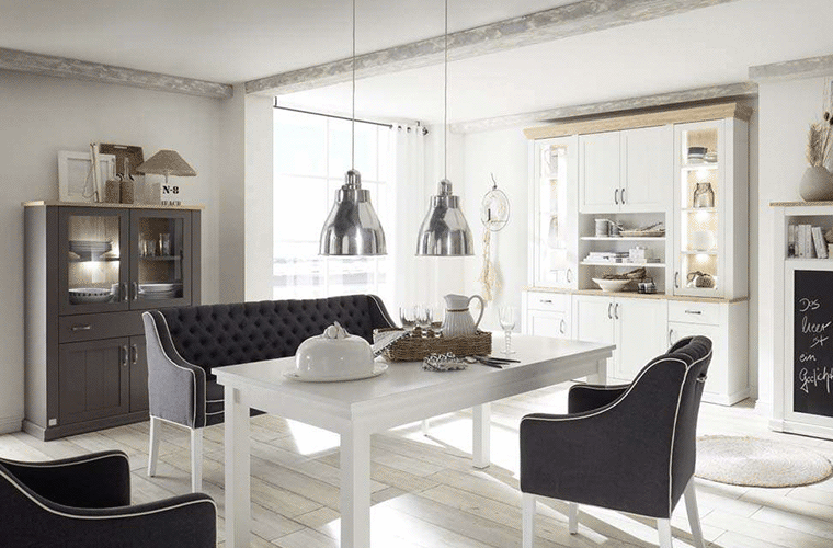 Essbereich im Landhausstil mit weißem Tisch, Vitrinenschränken und dunklen Polstermöbeln der Marke Sylt Living.