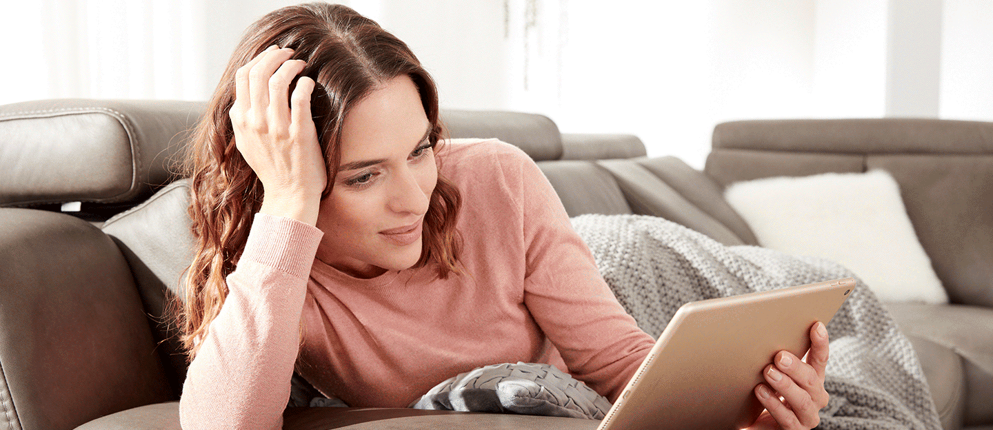 Frau liest entspannt und liegend ihr Prospekt auf einem braunen Sofa.