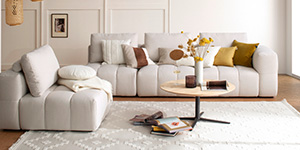 Eine helle Garnitur mit vielen Dekokissen steht in einem hellen Wohnzimmer.