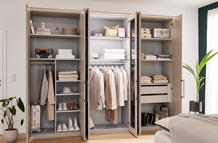 Offener Kleiderschrank in Beige steht in einem hellen Zimmer.