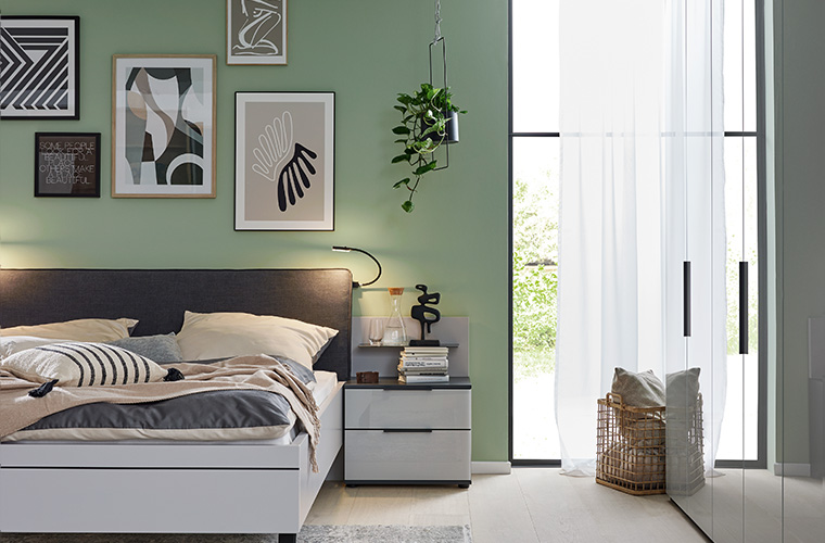 Ein graues Doppelbett und ein dazu passender Nachttisch stehen in einem grün gestrichenem Schlafzimmer.