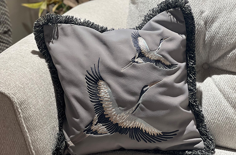 Dekoratives Kissen mit grauem Bezug und fransen auf einem Sofa.