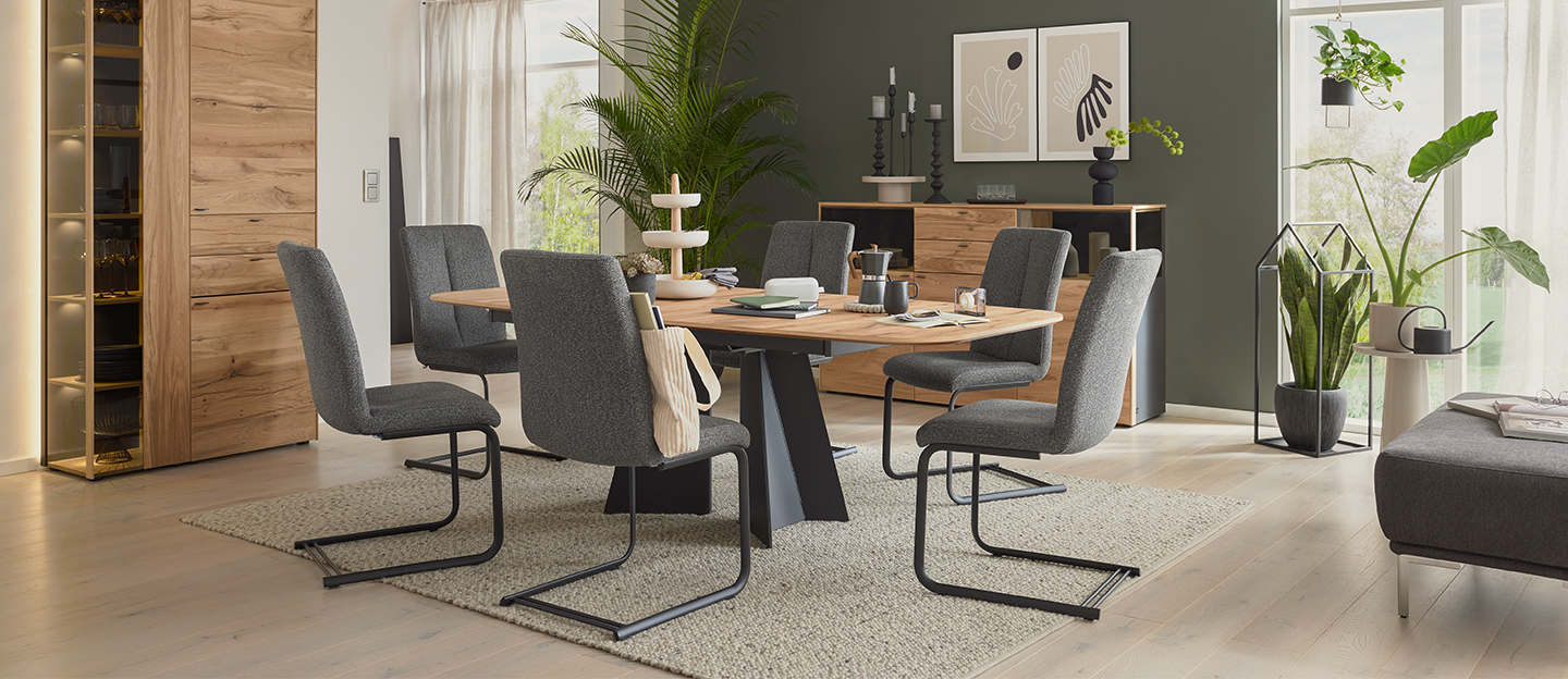 Esszimmer mit einem hellen Holztisch und grauen Stühlen, welches mit vielen grünen Pflanzen dekoriert ist.