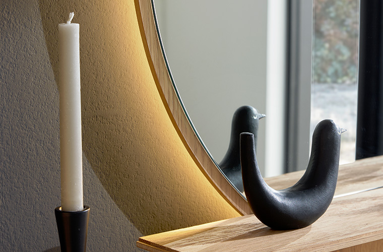 Detailaufnahme eines runden Spiegels mit Beleuchtung, der über einer Garderobe aus Holz hängt.