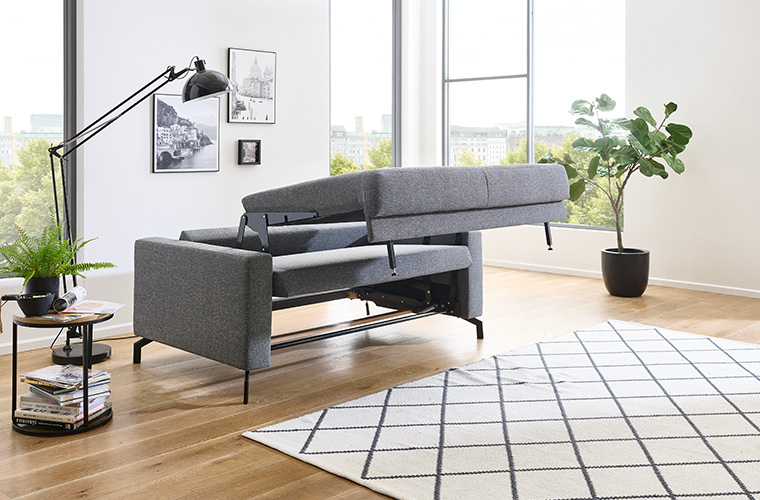 Modernes, graues Schlafsofa mit schwarzen Füßen in einem hellen Wohnzimmer.