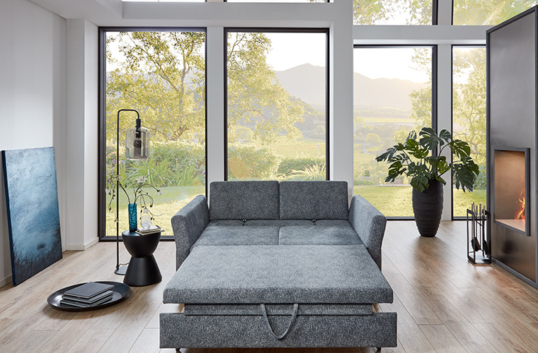 Ausgezogenes, graues Schlafsofa als Bett vor einer großen Fensterfront.