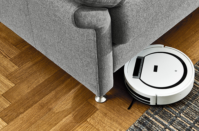 Ein runder Saugroboter reinigt den Boden unter einem grauen Sofa
