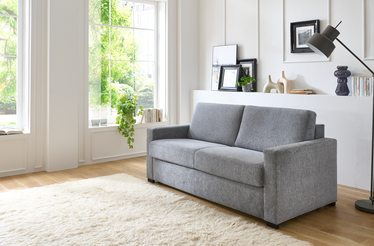 Schlafsofa „Aygo Comfort“ in Grau in einem hellen Wohnzimmer.