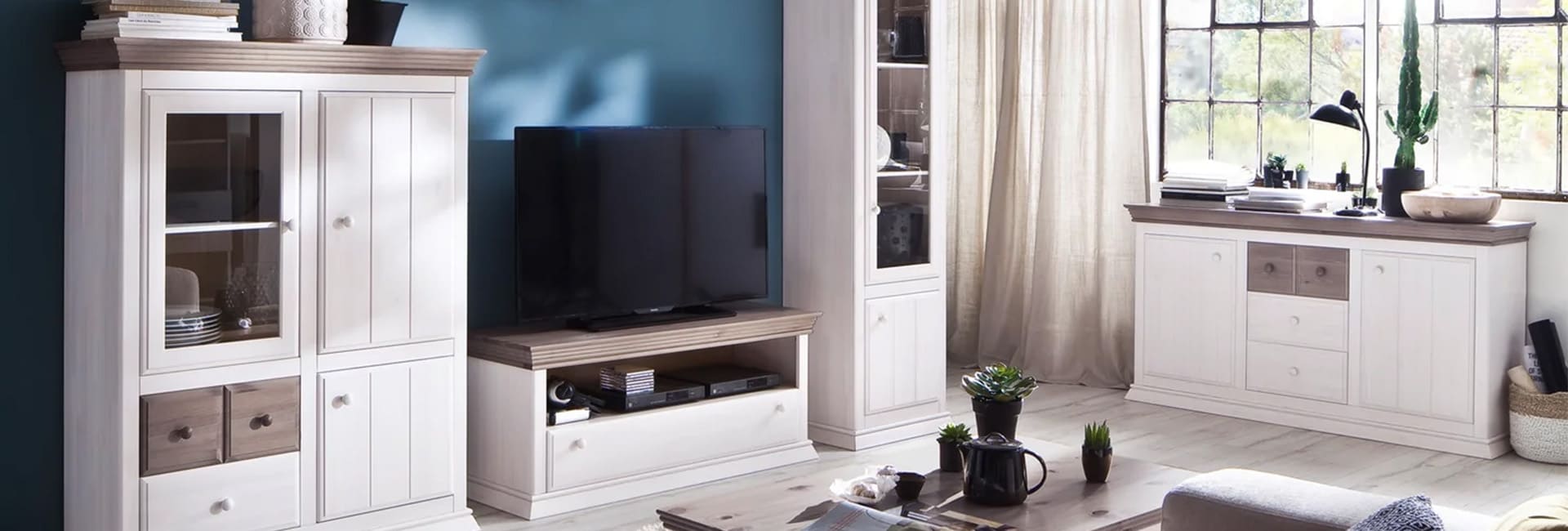 Heller Wohnraum im Landhausstil mit weißen Vitrinen- und Regalschränken der Marke Sylt-Living.