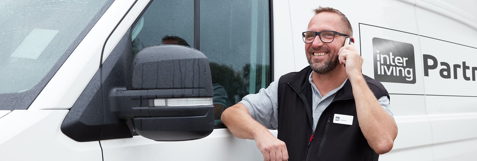 Monteur mit schwarzer Arbeitsweste telefoniert vor seinem weißen Servicefahrzeug.